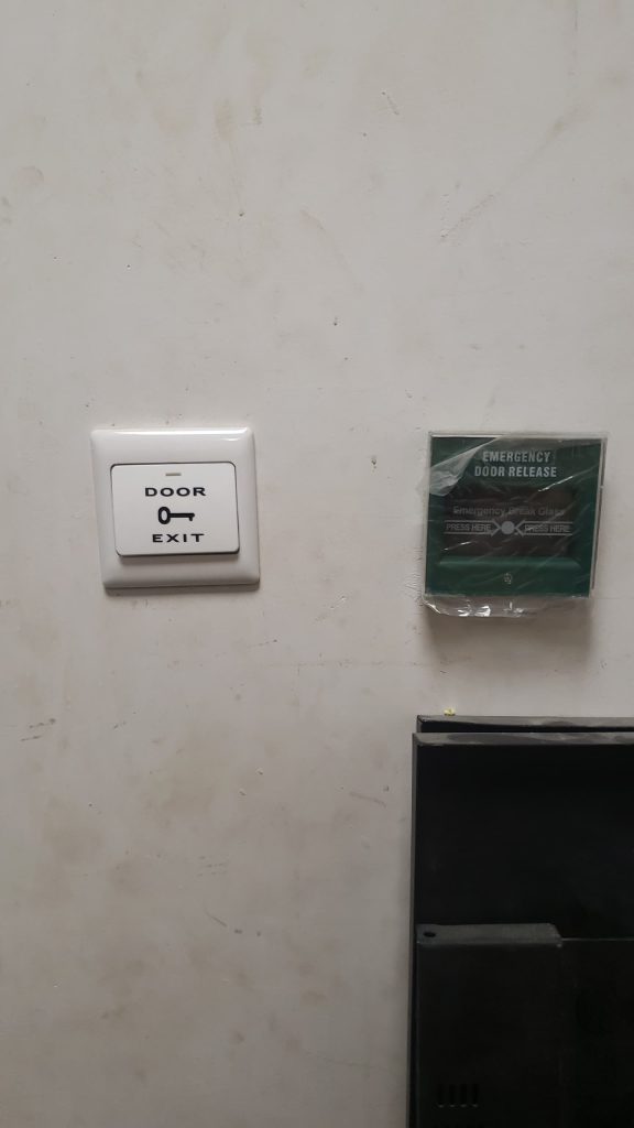 Wall-mounted door exit push button installed beside an emergency break glass unit for emergency door release.