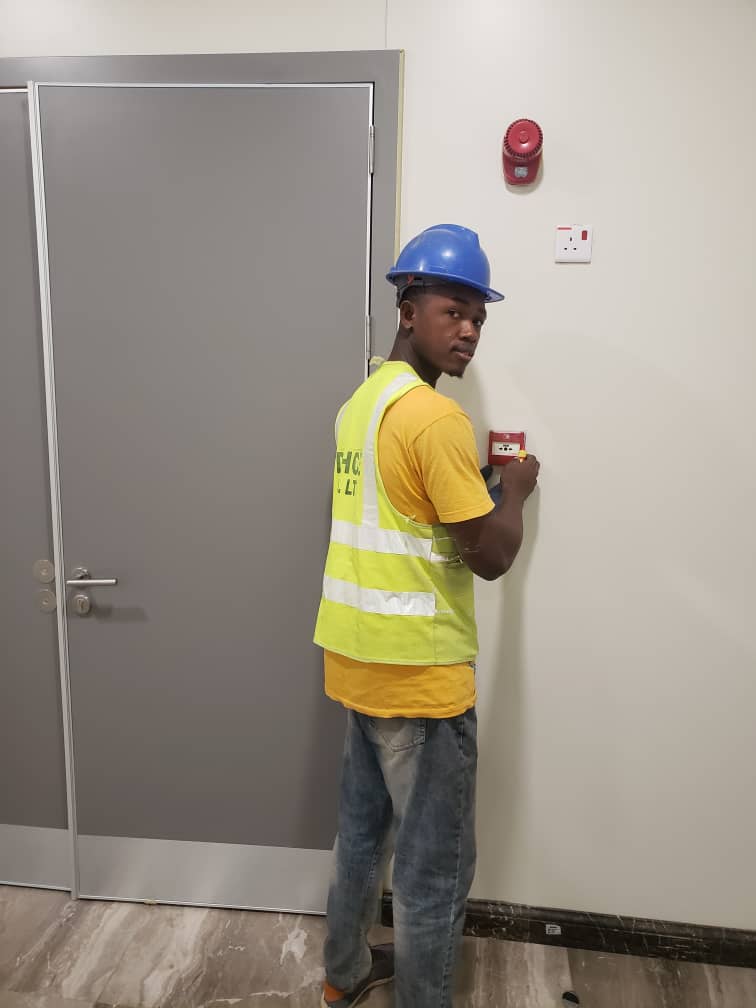Technician wearing safety helmet and reflective vest installing an emergency break glass unit on a wall beside a secured door.
