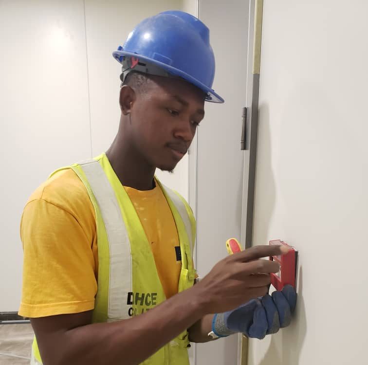 Technician wearing safety helmet and reflective vest installing an emergency break glass unit on a wall beside a secured door.